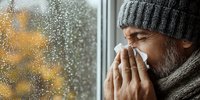 Middle-aged man with beard wearing winter hat and scarf blowing nose by rainy window with autumn leaves in background.