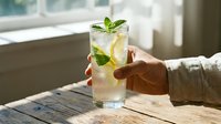 Refreshing mojito cocktail with mint and lime held by hand on rustic wooden table near bright window for summer beverage and bar menu concepts.