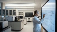 Female scientist in white lab coat writing chemical formulas on whiteboard in modern research laboratory with equipment and workstations in background.