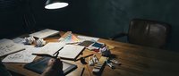 Creative workspace with scattered papers, art supplies and vintage camera on wooden desk under warm lamp light for artistic projects and design work.