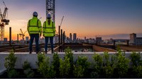 Two construction workers in high-visibility safety vests stand on a rooftop at sunset, overlooking an urban skyline with cranes and green plants, representing industrial development and workplace safety.