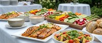 Outdoor catering buffet spread with grilled salmon, fresh salads, vegetables and bread rolls on white tablecloth for wedding reception or corporate event.