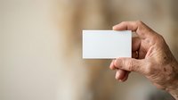 Mature man with wrinkled hands holding blank white business card against blurred neutral background, suitable for corporate branding and professional networking materials.