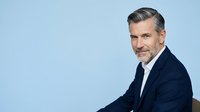 Middle-aged Caucasian male professional in dark blazer and white shirt against blue background. Corporate headshot for business, executive, leadership, and corporate identity use.