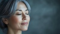Mature Asian woman with silver hair and closed eyes enjoying peaceful moment against dark background, conveying mindfulness and serenity.