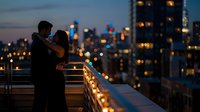 Romantic couple silhouette embracing on urban balcony at twilight with glowing city skyline and bokeh lights creating intimate evening atmosphere.