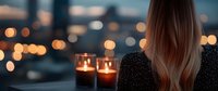 Young woman with long blonde hair looking at city lights from balcony with glowing candles creating romantic evening atmosphere against blurred urban bokeh background.