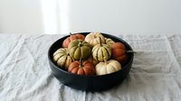 Colorful miniature pumpkins and gourds arranged in black bowl on white linen tablecloth for autumn harvest decoration and seasonal home styling.