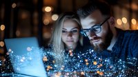Caucasian couple looking at glowing laptop screen with magical digital particles effect, young bearded man wearing glasses and blonde woman in dark room with bokeh.