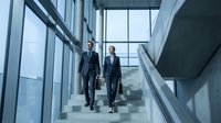 Two professional businesspeople walking down modern office stairs in corporate building with glass windows and contemporary architecture.