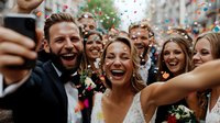 Joyful wedding celebration with bride, groom and friends taking selfie surrounded by colorful confetti shower during outdoor ceremony. Natural candid moment captures pure happiness.