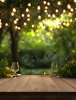 Empty wine glasses on wooden table in garden setting with string lights and green foliage creating romantic outdoor dining atmosphere. Bokeh lighting effect adds warmth.