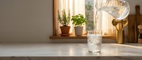 Fresh water being poured from glass pitcher into tumbler on kitchen counter with herbs and warm sunlight streaming through window.