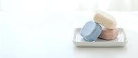 Stacked handmade soap bars in pastel blue, pink, and cream on white ceramic tray against neutral background. Spa and wellness product photography.