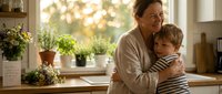 Loving grandmother embracing young grandson in warm kitchen setting with natural sunlight streaming through windows creating cozy family moment.