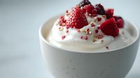 Fresh strawberries and cranberries on top of smooth whipped cream in white ceramic bowl, garnished with pink peppercorns against light background.