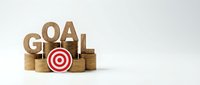 Financial goal achievement concept with wooden letters spelling GOAL, stacks of golden coins and red target symbol on white background for business planning.