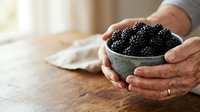 Mature man with light skin tone holding a ceramic bowl filled with fresh black caviar on wooden table surface with natural window lighting.
