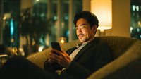 Young Asian businessman relaxing on couch using smartphone at home in evening with warm lighting and city view through windows.