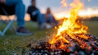 Glowing campfire with burning logs and sparks at sunset, friends relaxing in camping chairs in background, creating cozy outdoor atmosphere for summer adventures.