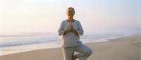 Mature woman practicing yoga in tree pose on sandy beach at sunrise, wearing casual athletic clothing, promoting wellness and mindfulness outdoors.