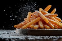 Crispy golden french fries sprinkled with salt on dark wooden board against black background with dramatic lighting and motion of falling salt particles.