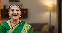 Mature woman of South Asian ethnicity wearing traditional green saree with gold embroidery, smiling warmly indoors. Suitable for cultural representation, family, lifestyle, and diversity stock content.