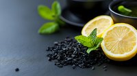 Fresh lemon slices with mint leaves and scattered black tea leaves on dark slate background, moody food photography composition for beverage concept.
