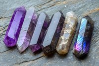 Raw crystal points arranged in gradient from purple amethyst through clear quartz to labradorite on gray stone background, showing natural mineral formation and healing stones.
