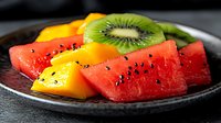 Fresh tropical fruit slices arranged on dark plate including watermelon, mango and kiwi garnished with black seeds, close up view of healthy summer dessert.