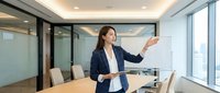 Asian female professional in dark blazer presenting business strategy in modern high-rise conference room with city skyline view, suitable for corporate training and leadership content.