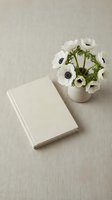 Minimalist workspace with blank hardcover book and white anemone flowers in ceramic vase on neutral linen background for creative projects and branding.