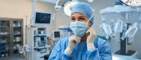 Female surgeon in blue scrubs and surgical mask standing confidently in modern operating room with medical equipment and surgical lights in background.