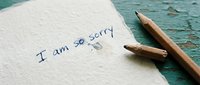 Handwritten apology note reading I am so sorry on textured paper beside a pencil on a rustic teal wooden surface.