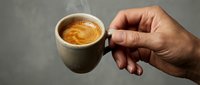Unrecognizable person holding ceramic mug with hot espresso coffee and latte art foam against neutral gray background for beverage and lifestyle use.