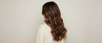 Young woman with long wavy brown hair wearing white sweater from behind against neutral beige background for fashion and beauty concepts.