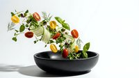 Fresh mixed salad ingredients with cherry tomatoes and herbs falling into black bowl on white background for healthy cooking and meal preparation.