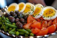Fresh Nicoise salad ingredients arranged on plate with halved boiled eggs, black olives, green beans, and cherry tomatoes garnished with herbs and seasonings.