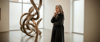 Middle-aged woman with blonde hair, Caucasian ethnicity, wearing black formal attire, observing abstract wooden sculpture in contemporary art gallery or museum space with natural light.