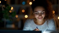 Young African American woman studying at night with glowing bokeh lights, smiling while writing in notebook, cozy evening atmosphere at home.