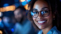 Young African American businesswoman with glasses smiling confidently in modern office environment with bokeh city lights background at night.