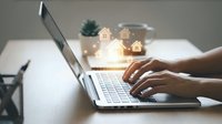 Real estate online search concept with hands typing on laptop and glowing house icons floating above keyboard for property investment and home buying.
