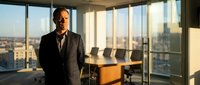 Professional middle-aged Caucasian businessman in dark suit standing confidently in modern office boardroom with panoramic city skyline view during golden hour.