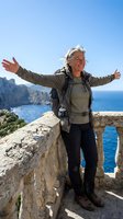 Happy senior woman hiker with arms outstretched celebrating on coastal cliff overlooking blue ocean waters during outdoor adventure vacation.