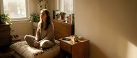 Young woman sitting cross-legged on bed in cozy bedroom with warm sunlight, plants and wooden furniture creating peaceful morning atmosphere.