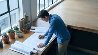 Young Asian male architect reviewing construction blueprints at modern office desk with plants and natural lighting for professional design work.