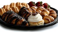 Fresh assorted croissants and pastries on black platter with chocolate drizzle, whipped cream filling and strawberries, arranged for catering or bakery display.