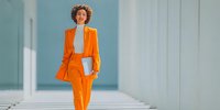 Confident young woman in vibrant orange business suit walking through modern minimalist hallway with white laptop, representing professional success and contemporary style.