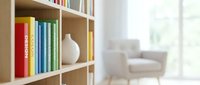 Modern wooden bookshelf with colorful books and white ceramic vase in bright living room interior with blurred armchair background for home decor concepts.
