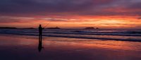 Silhouette of fisherman casting line at sunset beach with dramatic purple and orange sky reflecting on wet sand and rolling waves.
